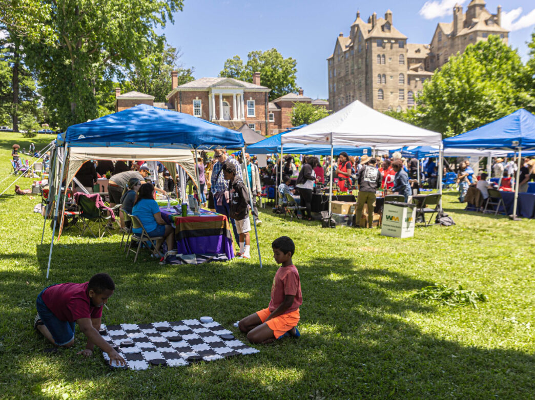 Two kids sit in the grass playing checkers on a large checkers board while vendor tents are set up behind them at the Mercer Museum and Fonthill Castle for a Juneteenth celebration.