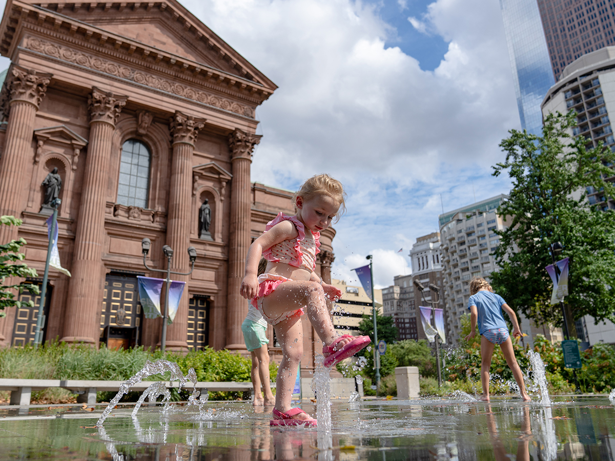 Een kind in een roze zwempak en roze sandalen speelt in een fontein in Sister Cities Park in Philadelphia.
