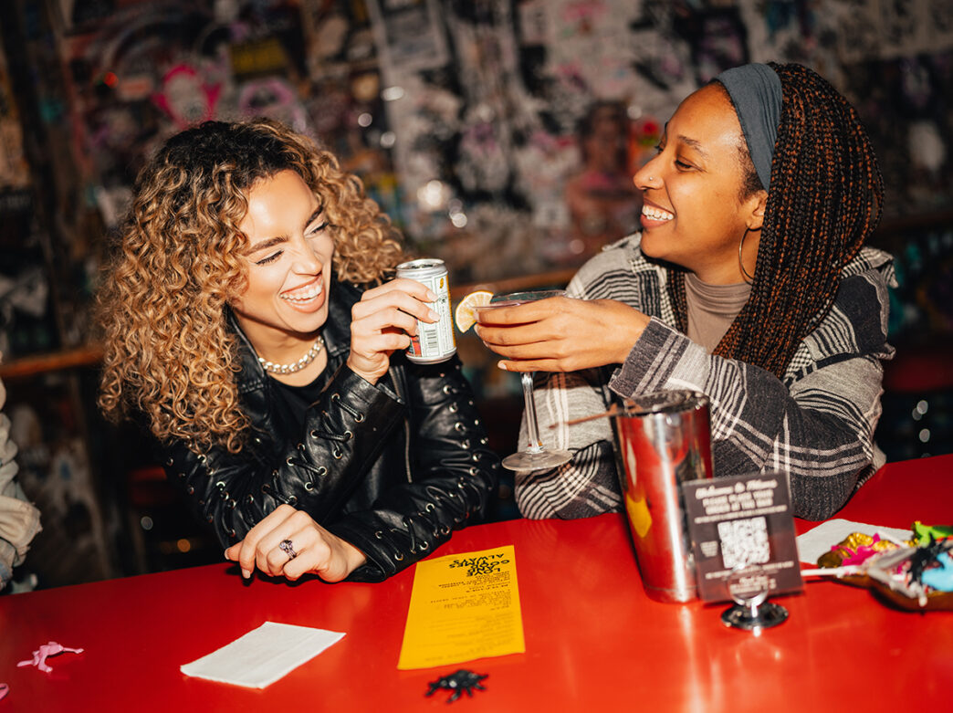 Two people sit at a table with a red tabletop and cheers their drinks while laughing at Tattooed Mom in Philadelphia.