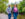 A woman wearing a pink shirt, leather jacket and jeans walks forward along a gravel trail while holding hands with a man behind her wearing a gray sweater and jeans.