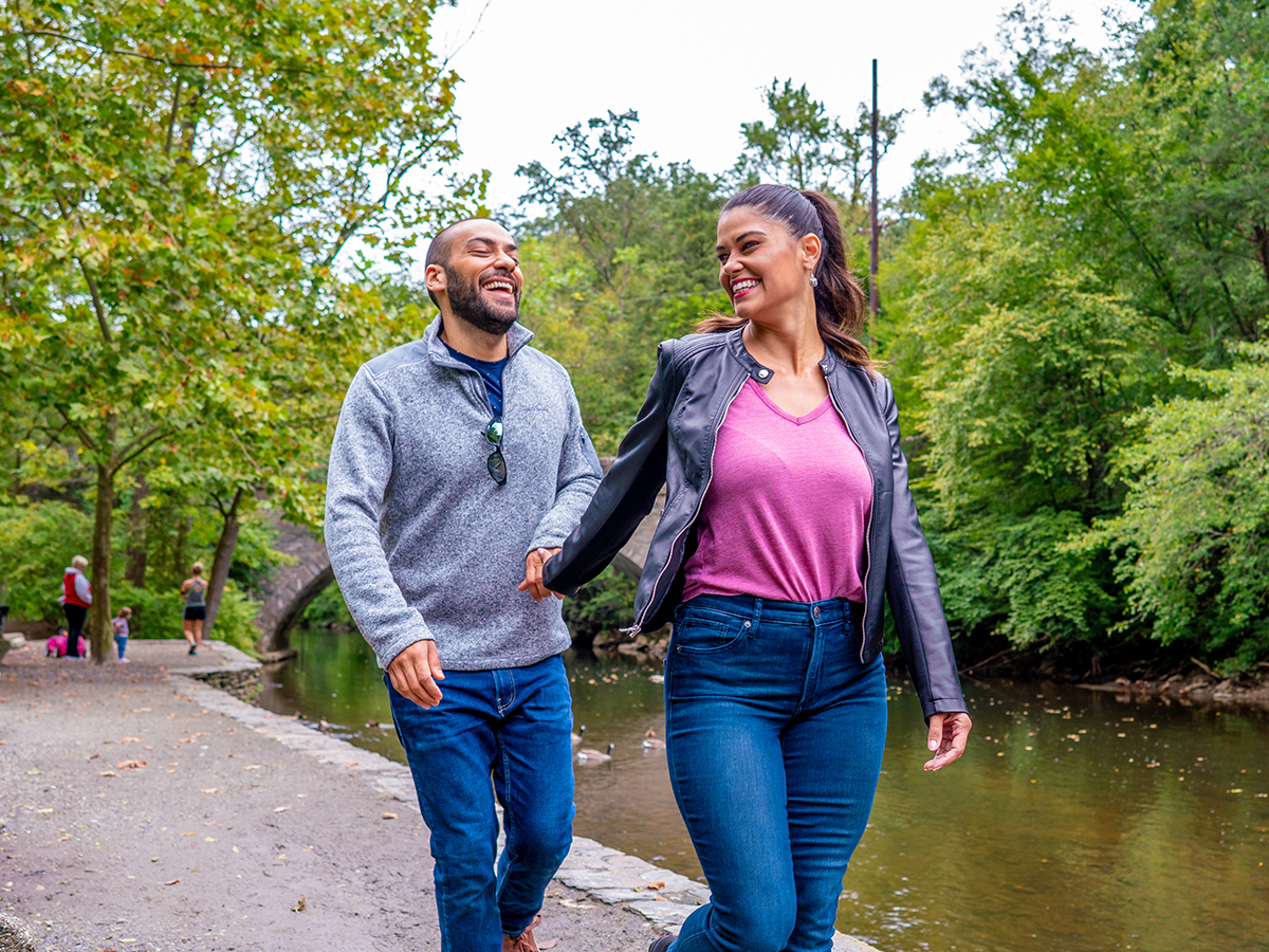 A woman wearing a pink shirt, leather jacket and jeans walks forward along a gravel trail while holding hands with a man behind her wearing a gray sweater and jeans.