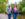 A woman wearing a pink shirt, leather jacket and jeans walks forward along a gravel trail while holding hands with a man behind her wearing a gray sweater and jeans.