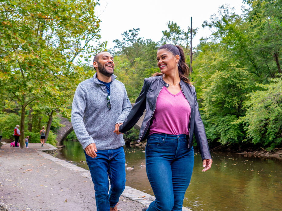 A woman wearing a pink shirt, leather jacket and jeans walks forward along a gravel trail while holding hands with a man behind her wearing a gray sweater and jeans.