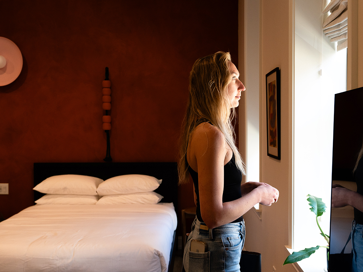 Uma pessoa olha pela janela enquanto está em um quarto de hotel. Uma cama com roupa de cama branca está ao fundo.