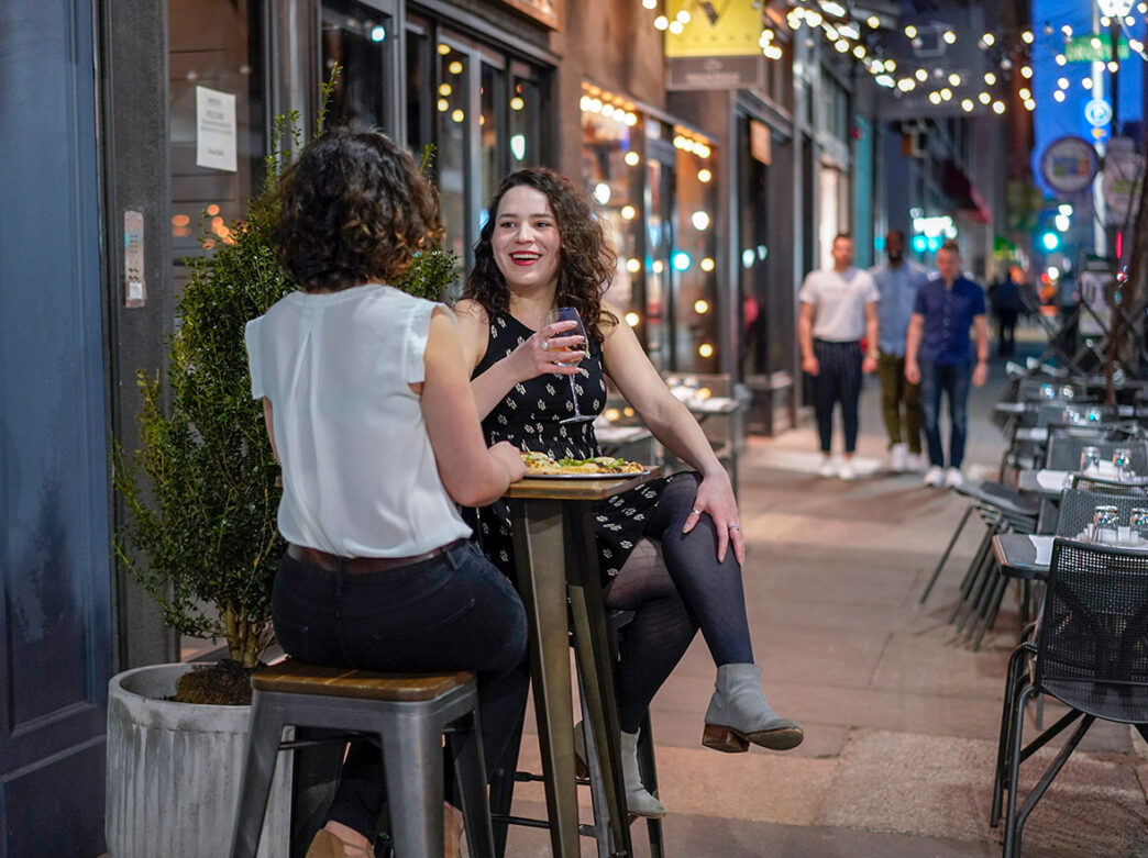Two people dine outside at a high top table at Barbuzzo. One person holds a wine glass in their hands.