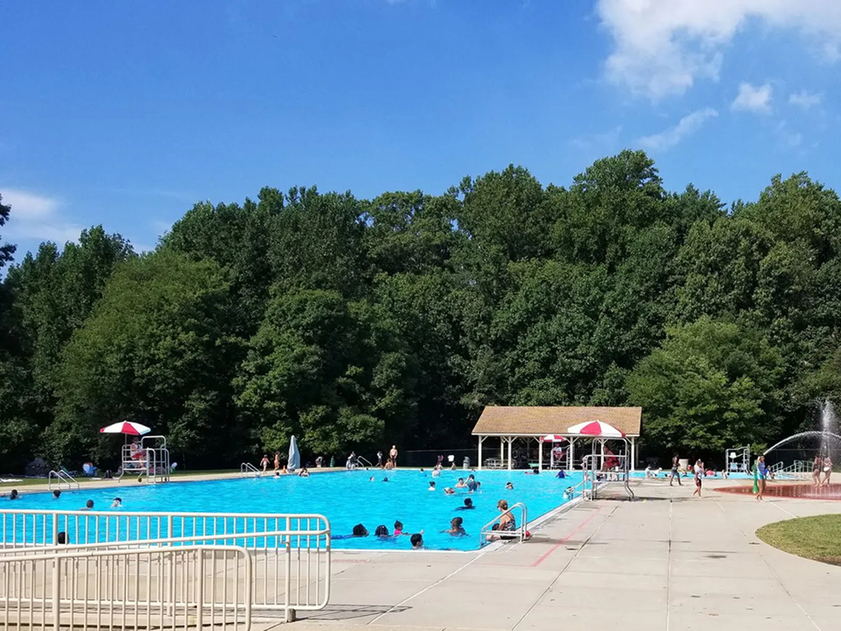 People swim in a large outdoor swimming pool with lifeguard stands and a pavilion near the pool.