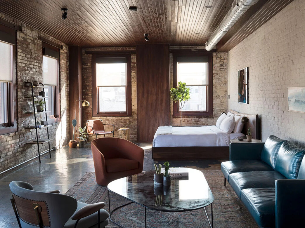 A hotel room furnished with a king bed and a seating area with exposed brick walls, concrete floors and four windows.