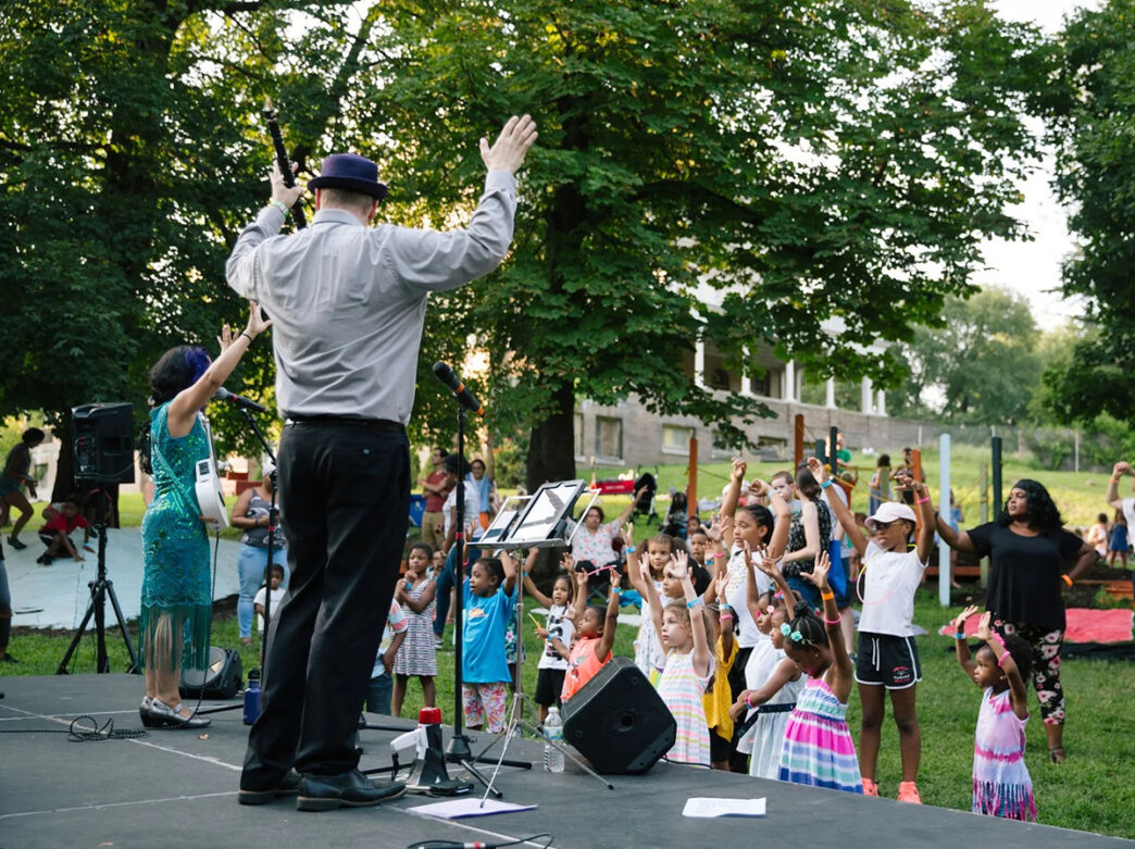 A group of children face a stage with their hands up in the air while musicians perform on stage during Kidchella.