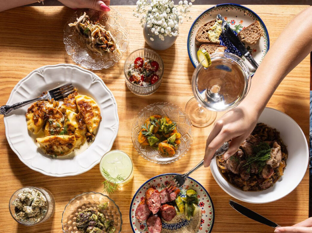 An aerial image of a person using a fork to pick up Kielbasa from a plate on a wooden table. Several other menu items, including pierogi and sourdough bread, are displayed as well.