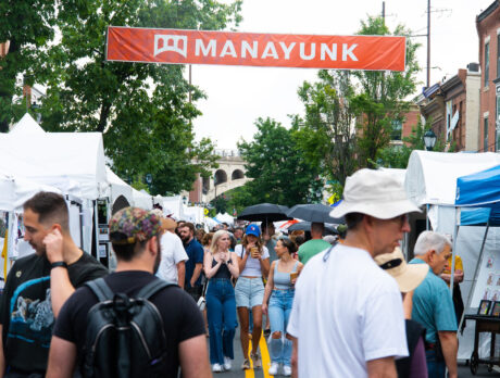 People walk through the crowd and a street lined with vender tents during the Manayunk Arts Festival. An orange banner with Manayunk in white lettering hangs above the street.