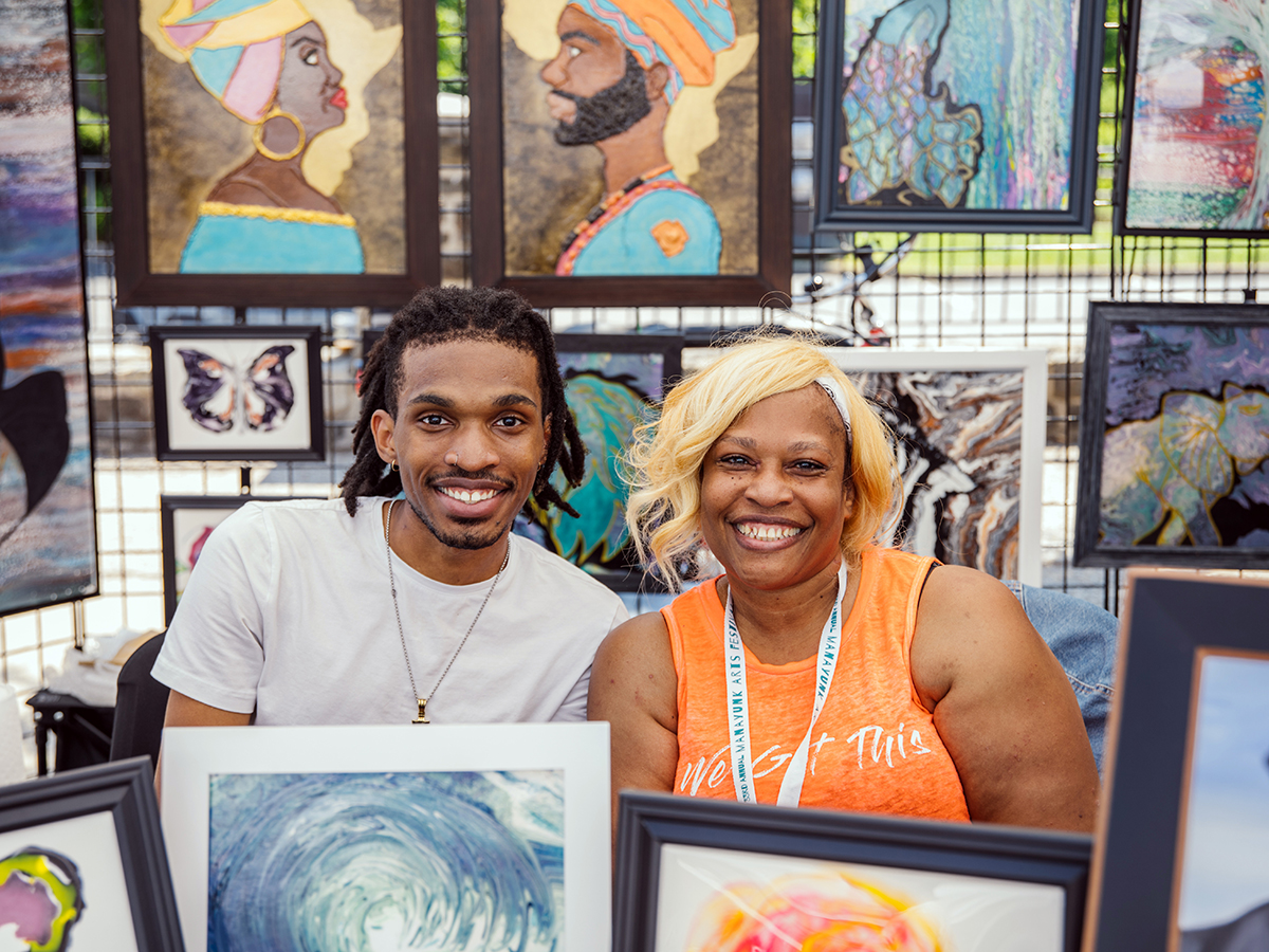 Two people sit a in a vendor tent displaying paintings for sale at the Manayunk Arts Festival.