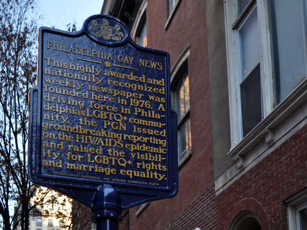 A navy historical marker with gold text is displayed in front of a red brick building.