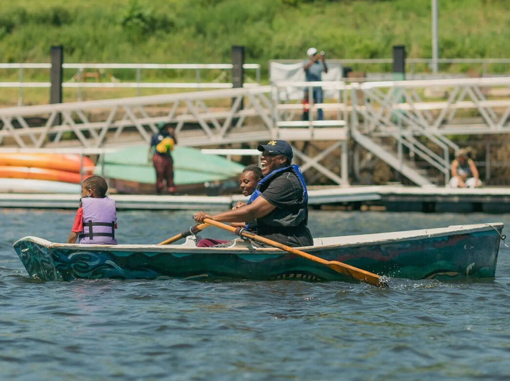 A person and two children wearing life vests row a row boat in a body of water near a dock.