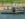 A person and two children wearing life vests row a row boat in a body of water near a dock.