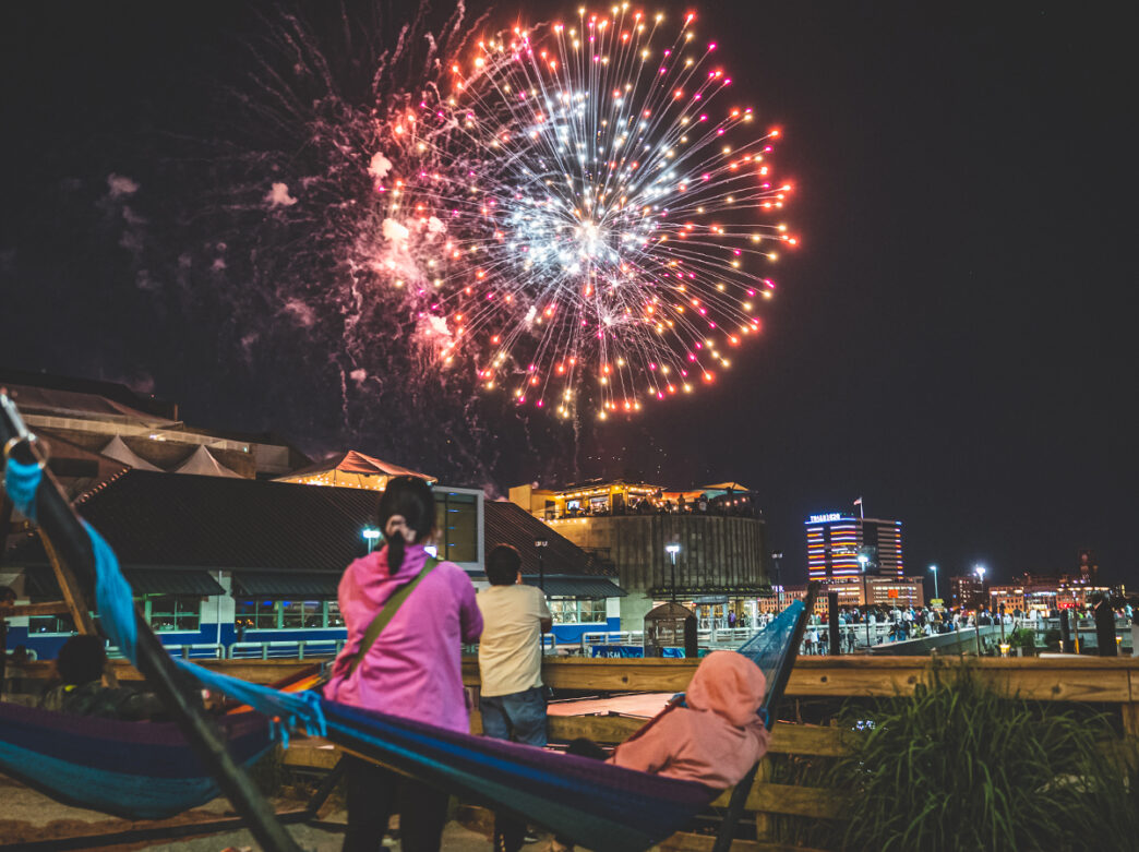 A red and pink firework explodes in the sky while people relaxing in hammocks at Spruce Street Harbor Park watch the firework show.