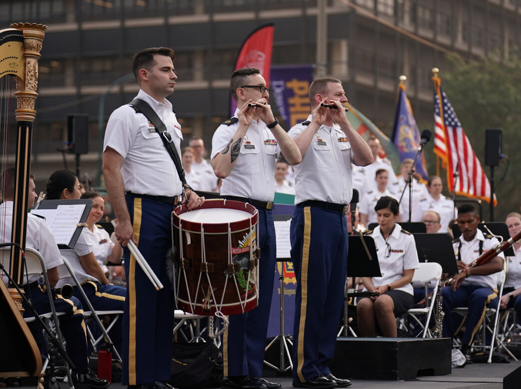 Three US Army personnel wearing white button down shirts and navy pants stand on stage and play instruments with the US Army Field Band for Wawa Welcome America.