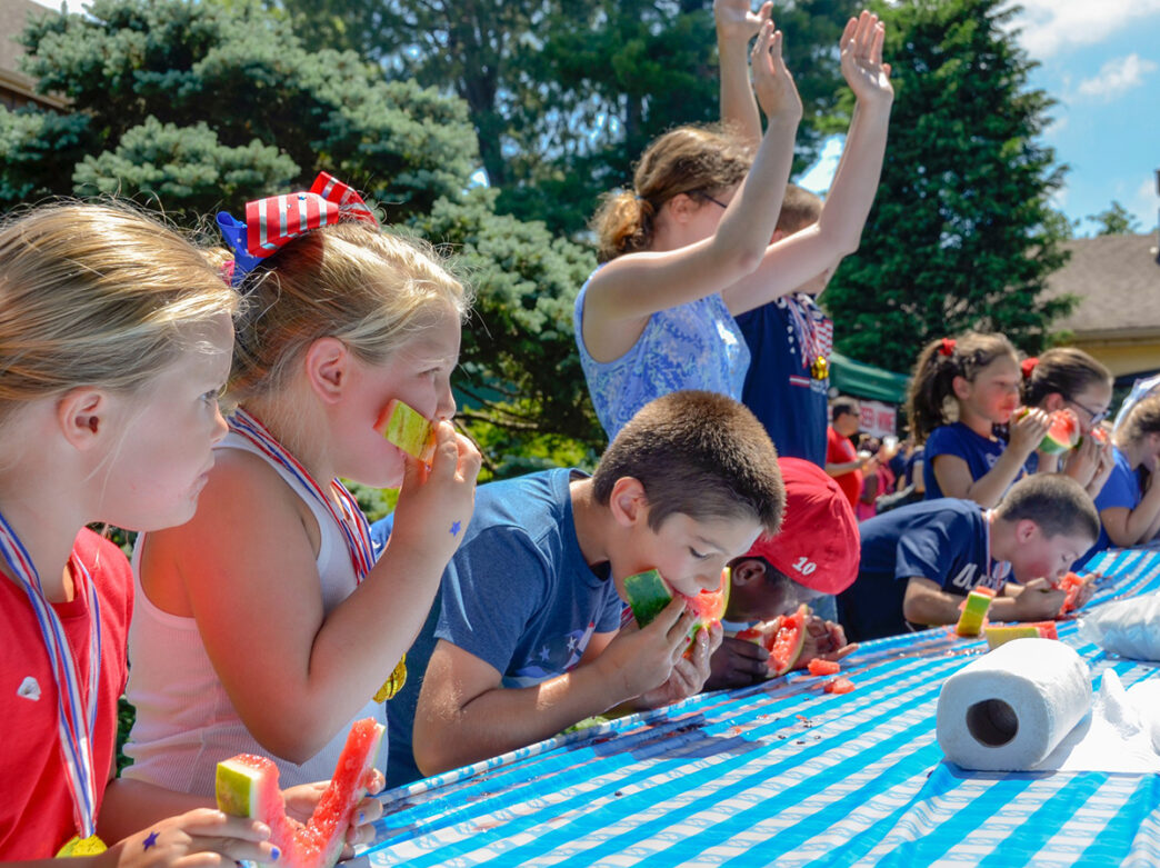 Children wearing red, white and blue shirts line up at table and eat watermelon during a watermelon eating contest at Peddler's Village.