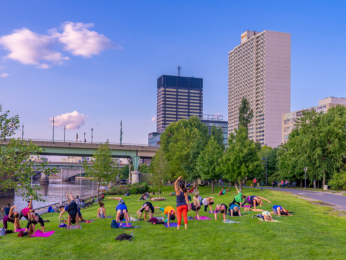 A group of people do yoga on the grass between the Schuylkill River and Schuylkill River Trail while an instructor leads the group.