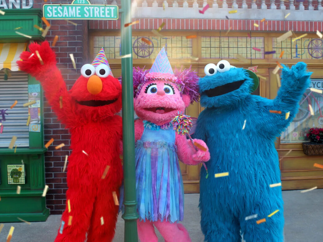 Elmo, Abby and the Cookie Monster pose for a photo together while wearing party hats and confetti drops from the sky.