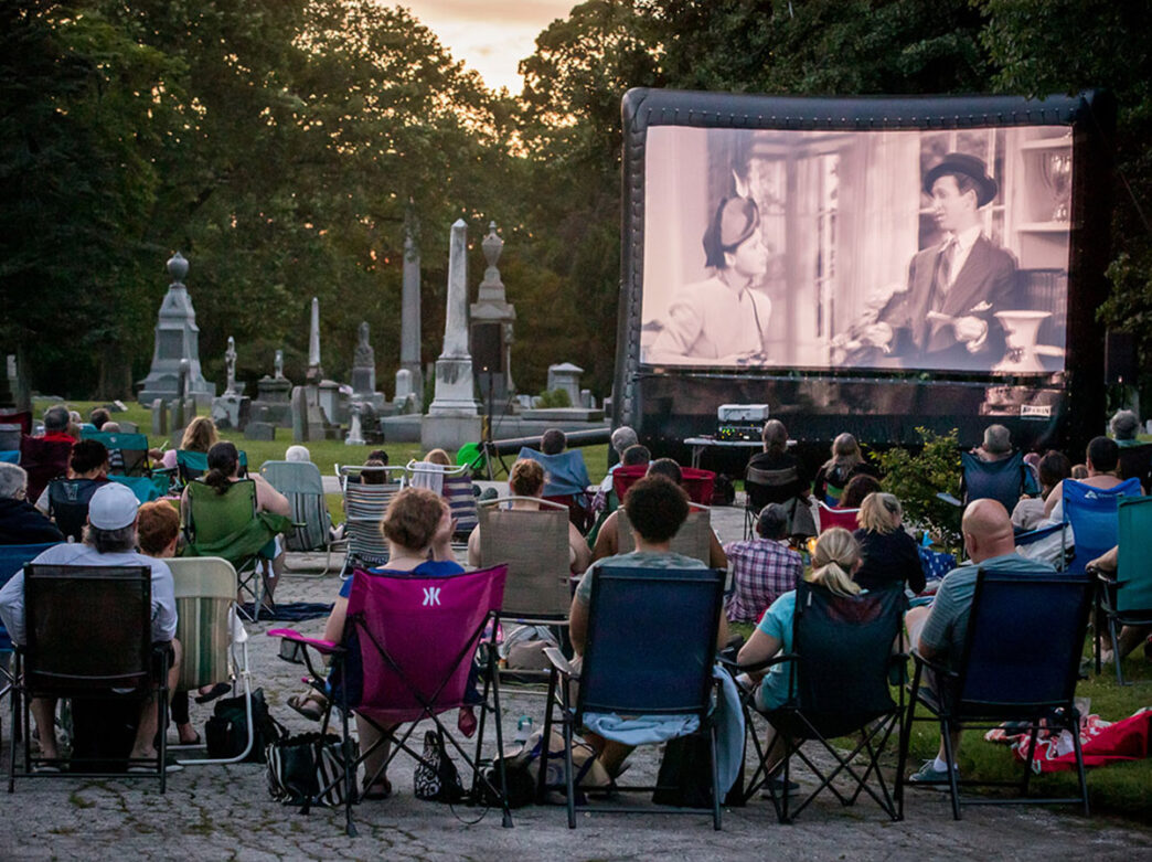 A crowd of moviegoers sitting on outdoor lounge chairs watch a movie amongst the graves at Laurel Hill Cemetery.