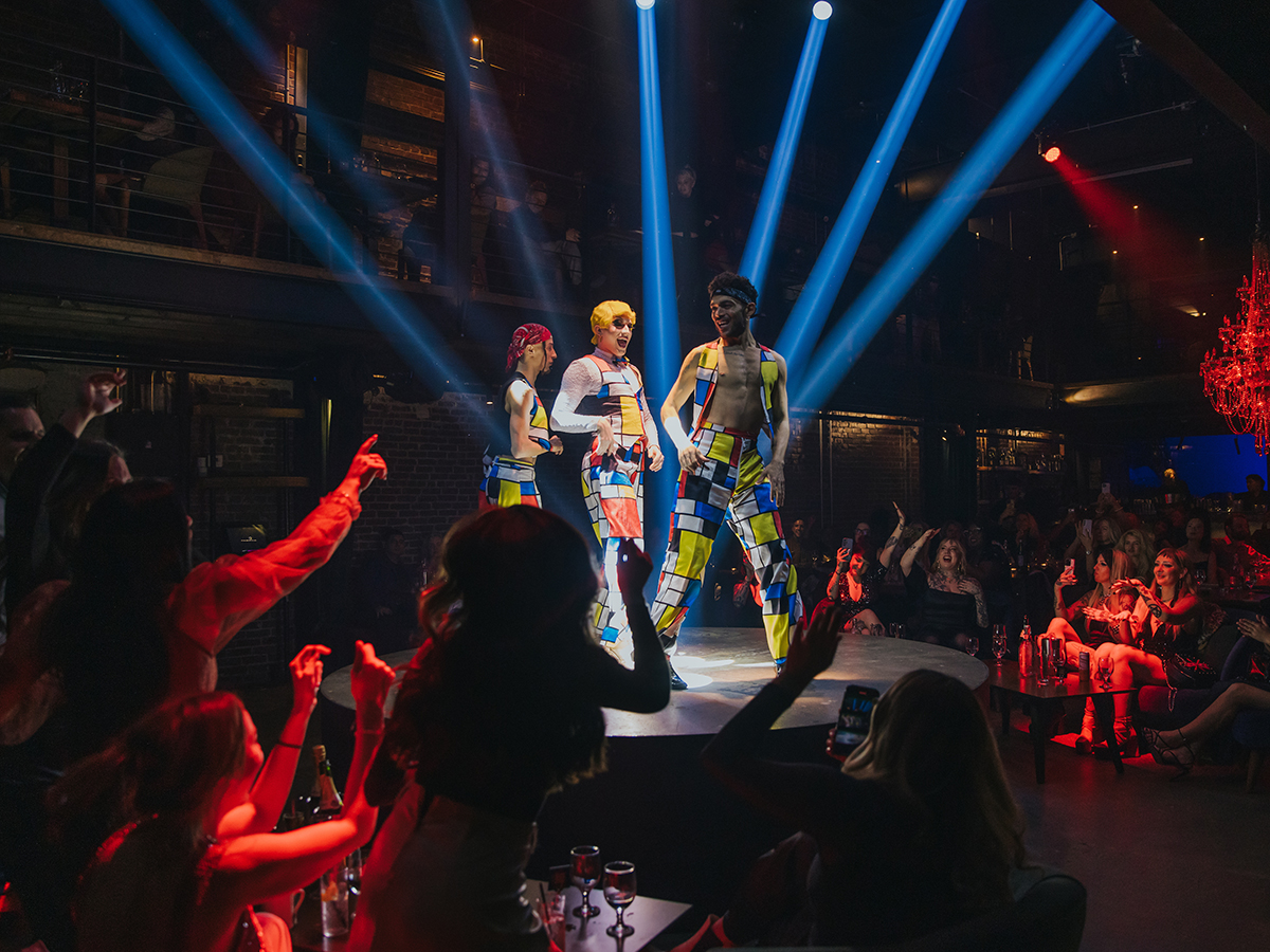 Three performers wearing colorful pants dance on stage at Fabrika while the crowd cheers and takes photos.