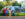 Four kids line up at a metal fence to watch a fountain show in Franklin Square. A carousel and green trees are in the background.