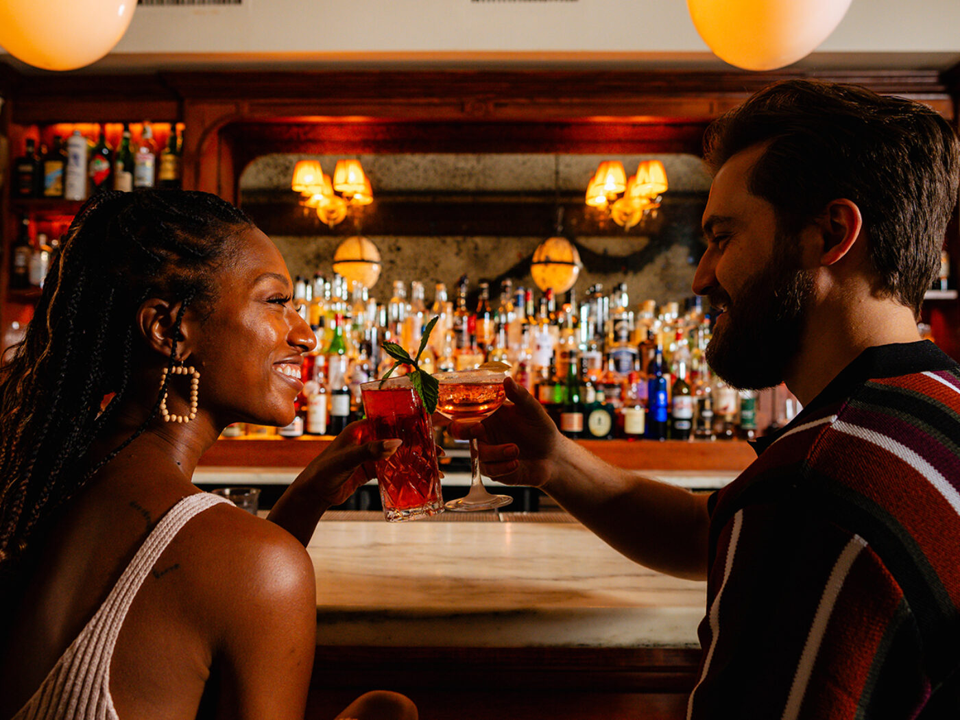 Two people sit at a bar and cheers each other with cocktails in their hands.
