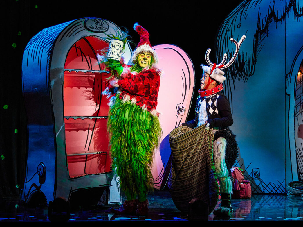 An actor dressed as the Grinch in a green furry outfit holds up a can taken out of a fridge. Another actor dressed as the dog, Max, holds a large bag and grimaces.
