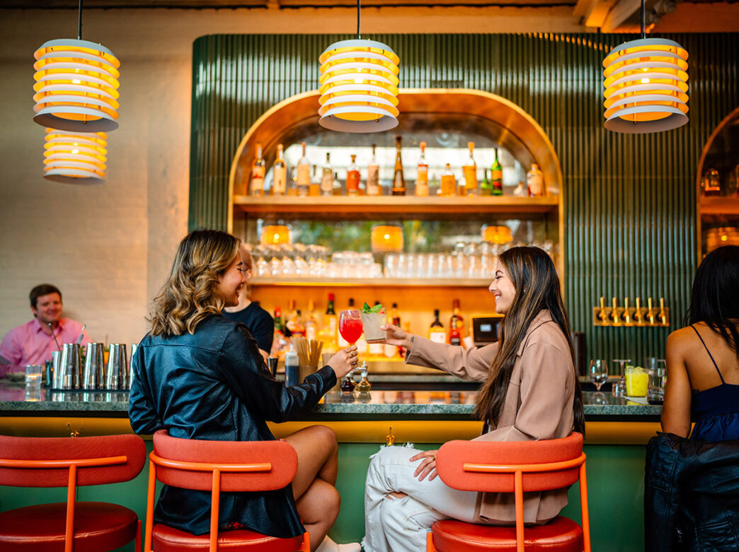 Dos mujeres jóvenes se sientan en la barra verde de Kalaya y brindan con sus cócteles.