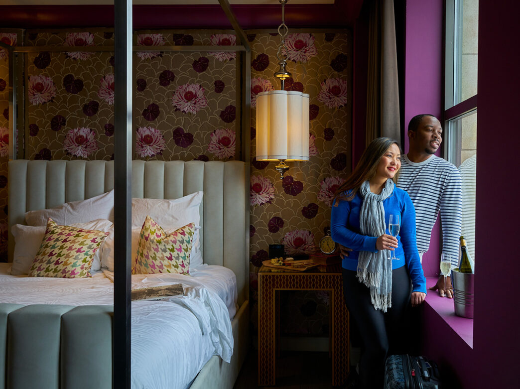 A couple looks out the window together while in a guest room at Kimpton Hotel Monaco in Philadelphia. The hotel room has a four poster bed and floral wallpaper.