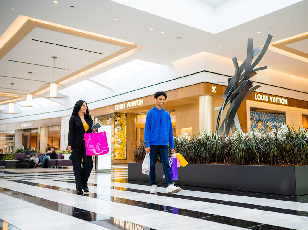 A casually dressed mother and son carrying colorful shopping bags walk in front of the Louis Vuitton store at King of Prussia Mall.
