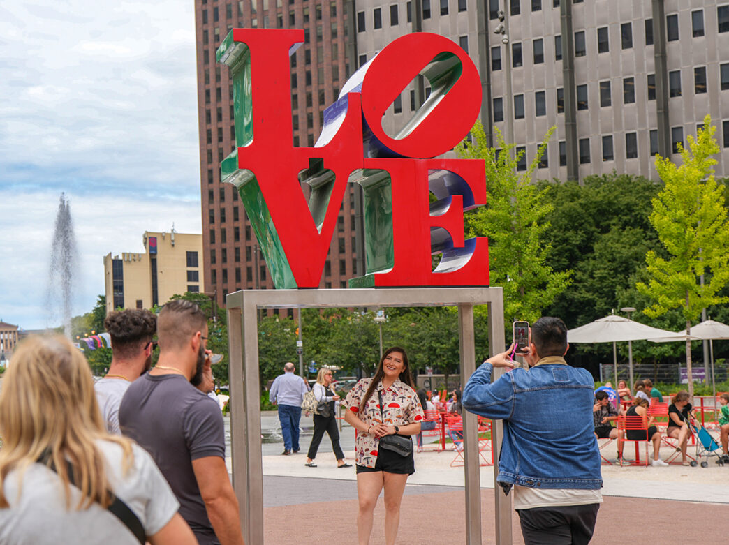 A person stands under the LOVE Statue while someone takes their photo with a cell phone. Other visitors wait in line to take their photo with the statue.