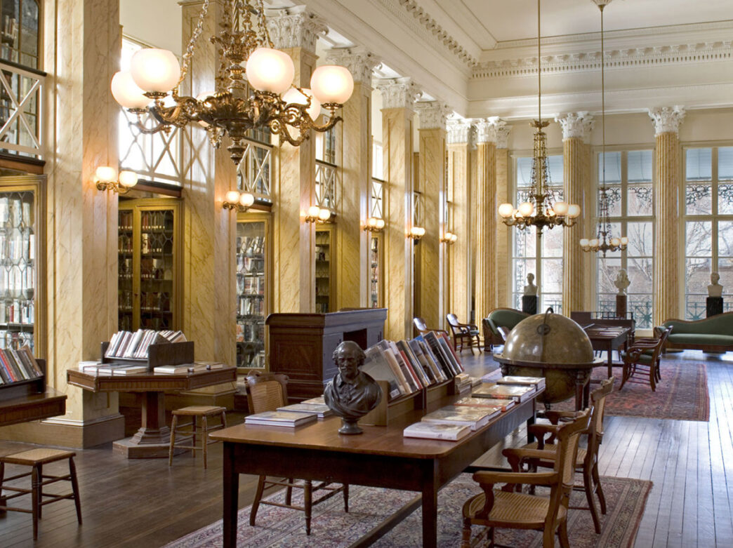 An elegant library decorated with gold light fixtures and wooden furniture displays books on tables and shelves.