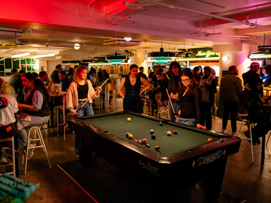 Cuatro mujeres, rodeadas de una multitud de comensales, juegan una partida de billar en el Middle Child Clubhouse.