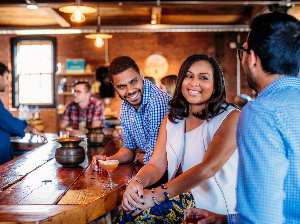 Una mujer vestida con chaleco blanco y falda azul estampada se sienta en la barra de la destilería New Liberty con dos hombres, ambos vestidos con camisas azules de cuadros.