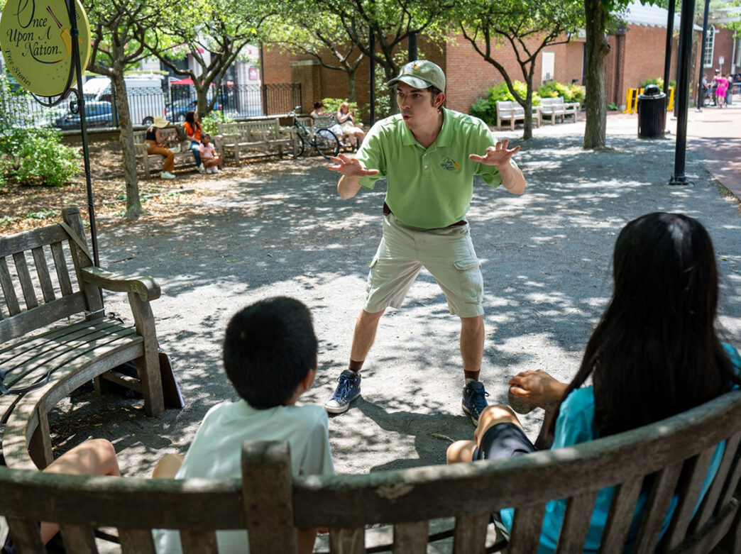 Three children sit on a bench outdoors while a storyteller performs in front of them.