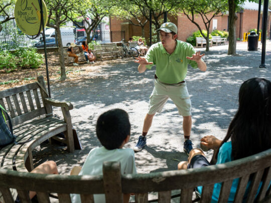 Three children sit on a bench outdoors while a storyteller performs in front of them.