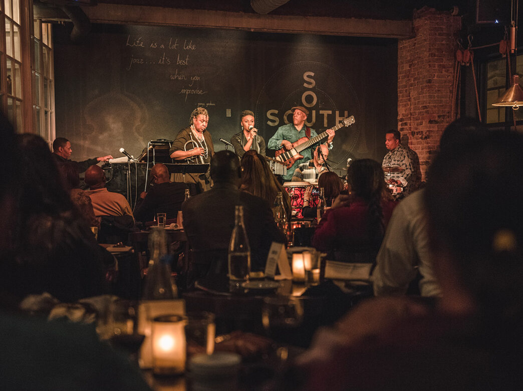 Una banda, formada por un cantante, un guitarrista, un pianista, un trombonista y un batería, sube al escenario ante un público abarrotado en SOUTH Jazz Kitchen.