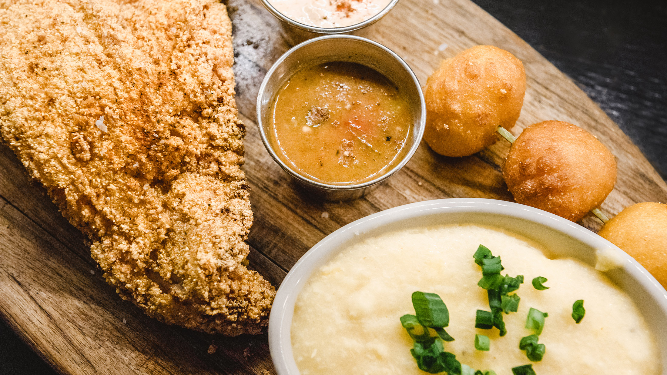 A delicious-looking assortment of catfish, mashed potatoes, hush puppies and dipping sauces sit on a wooden plate at SOUTH.