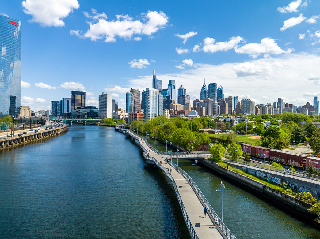 The Philadelphia skyline on a sunny day with the Schuylkill River and Schuylkill Banks Boardwalk in the foreground.
