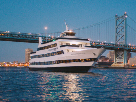 Un yate blanco de tres niveles navega de noche por el río Delaware con el Benjamin Franklin Bridge al fondo.