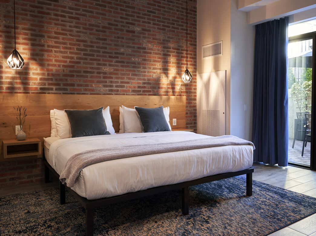 A king bed with white linen against a brick wall at The Maj in Philadelphia. Pendant lights hang on either side of the bed and sliding glass door looks out to a patio.