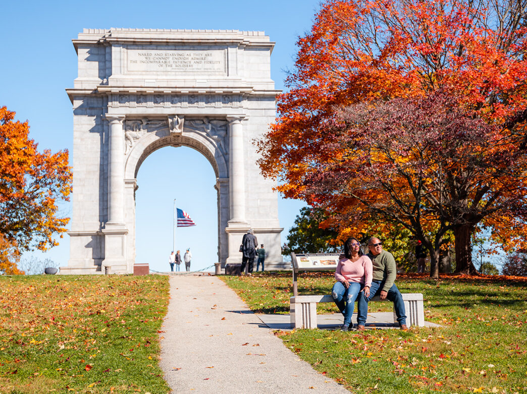 A couple sits on a bench in front of a large marble archway during the fall at Valley Forge National Park.
