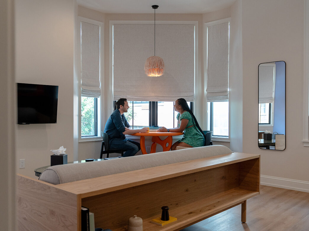 Two people sit facing each other at am orange breakfast table in a hotel room at Yowie. A full length mirror hangs on one wall and a TV hangs on another.