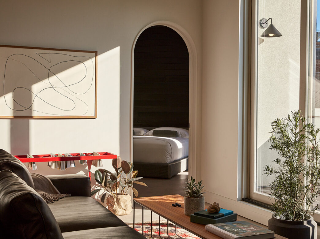 A living room with a couch, coffee table and potted plants looks into a bedroom through an arched doorway. Abstract art hangs on the wall and floor to ceiling windows let natural light into the room.