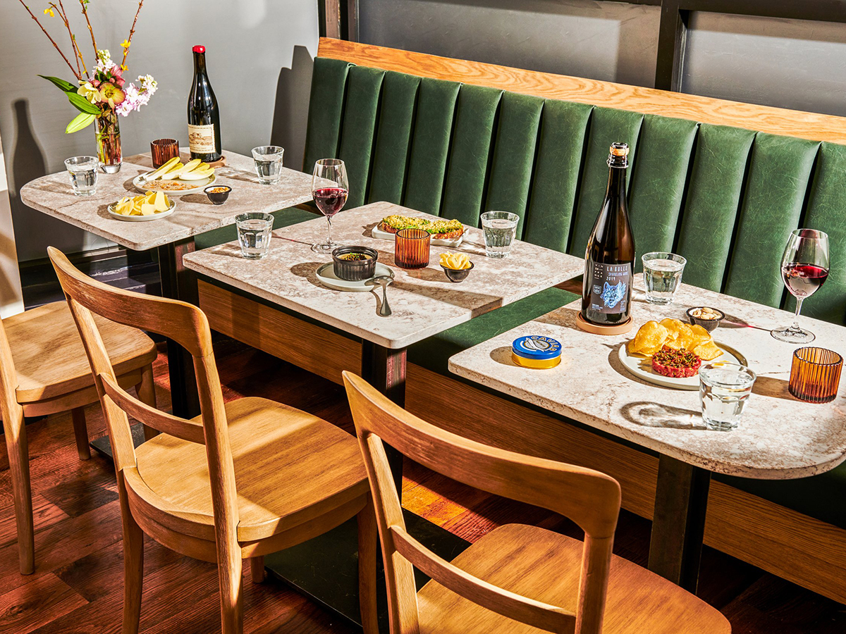 Three small tables line a green booth in a restaurant. Wine bottles, wine glasses and various snack items are displayed on the tables.