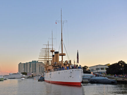 People stand on an old steel cruiser ship that is docked at the Delaware River Waterfront at sunset.
