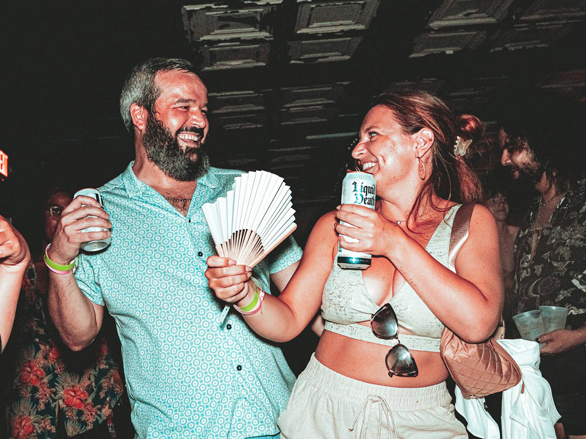 Two people look at each other and smile while dancing at Warehouse on Watts. They both hold beers in their hands and one person holds a paper fan.