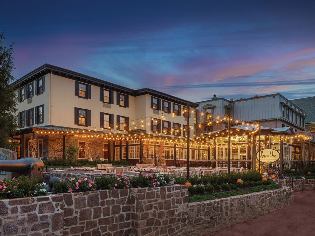 Exterior del Logan Inn por la noche con un cielo morado y azul oscuro. La propiedad está iluminada por luces de cuerda sobre un patio.