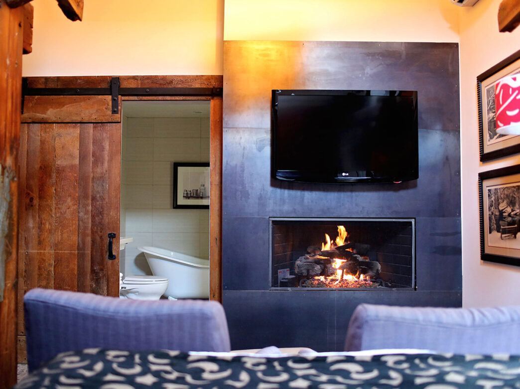 The camera faces a built in fireplace with a TV mounted above it and a reclaimed wood sliding door looking into a bathroom with a soaker tub.